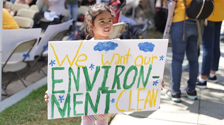 A small child holds a sign up at a protest against oil drilling in neighborhoods that reads "We want our ENVIRONMENT CLEAN."